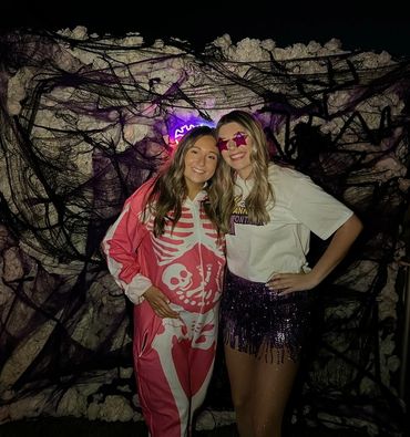 Two women in festive outfits pose in front of a Halloween-themed backdrop.