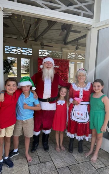 Santa and Mrs. Claus pose happily with four children in festive attire on a sunny day.