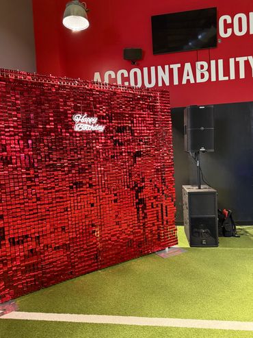 A red sequin backdrop with a 'Happy Birthday' neon sign and a speaker setup.