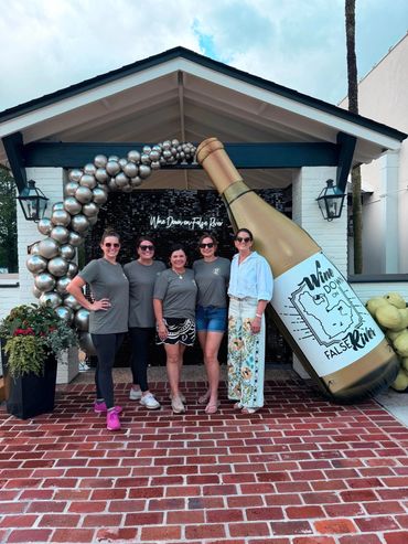 Five women posing with a large wine bottle prop and balloon arch.