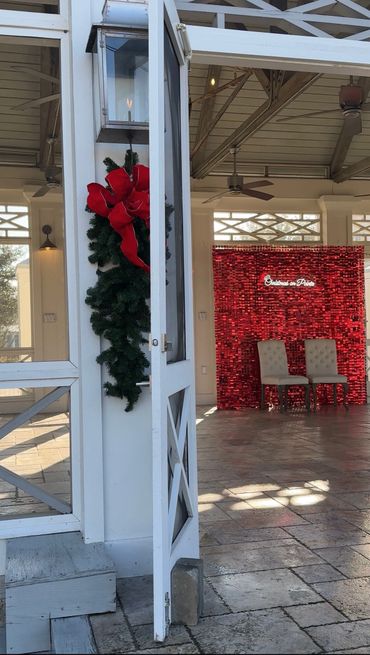 Open white door with Christmas wreath and red bow, festive red backdrop with chairs inside.