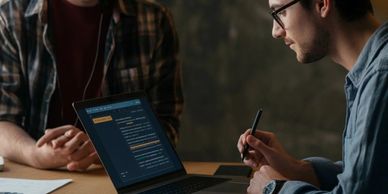 Two young men collaborating on coding with a laptop in a dimly lit room.