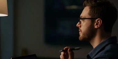 Man with glasses working late on a laptop, holding a pen thoughtfully.