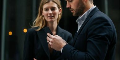 Two professionals in business attire, the man focused on his phone, the woman observing attentively.