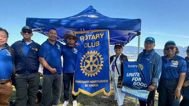 A group of Rotary Club members stand under a blue tent holding banners by the water.