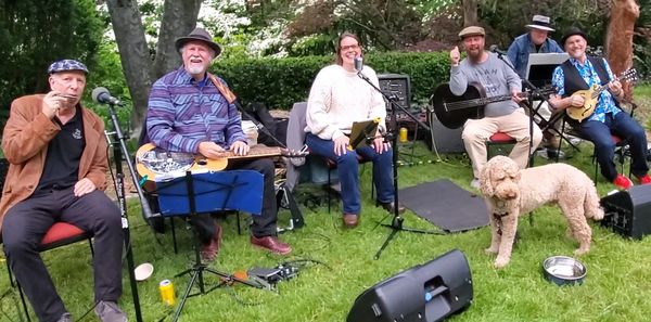 Group of musicians with instruments and a dog outdoors on grass.