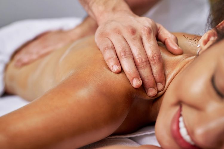Photo of woman smiling while getting back and shoulders massaged by a licensed therapist.