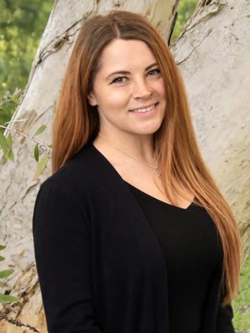 Smiling woman with long red hair standing outdoors by a tree.