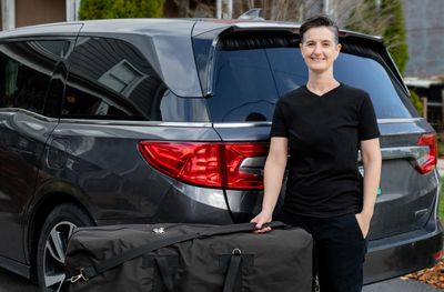 A photo of Meghan standing next to a vehicle holding strap of massage table in carrying case.