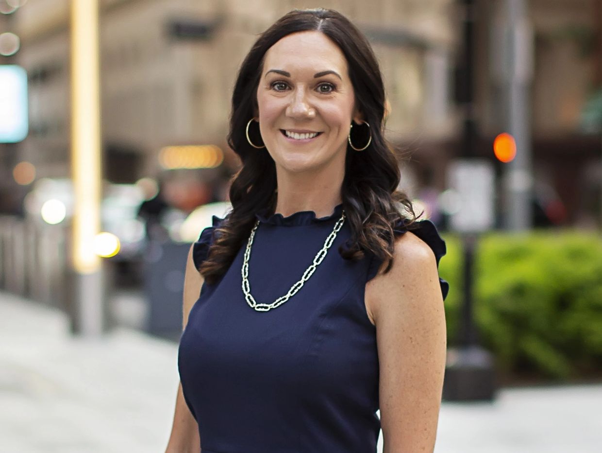 Smiling woman in a navy dress with hoop earrings and necklace in an urban setting.