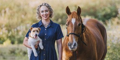 Woman in blue dress holding a dog and leading a horse in a nature setting.