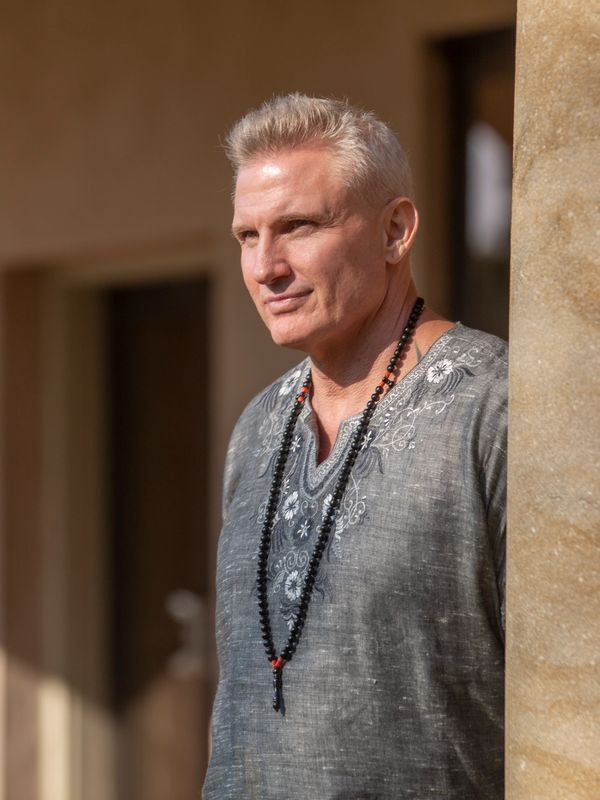Mature man in a gray embroidered shirt and beaded necklace stands thoughtfully by a pillar.