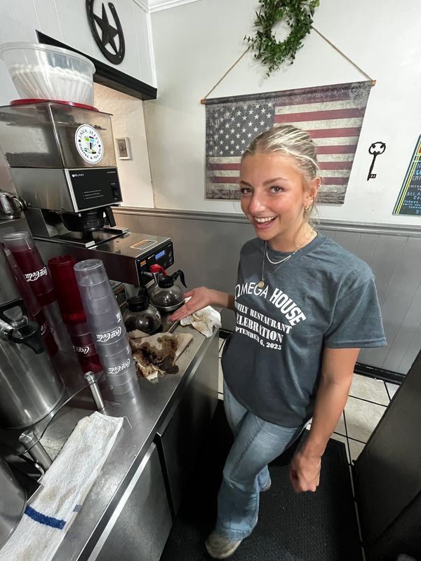 Smiling woman gestures at spilled coffee on a counter in a cozy cafe.