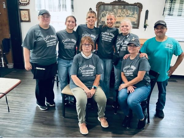 Group of eight people wearing matching Omega House celebration shirts indoors.