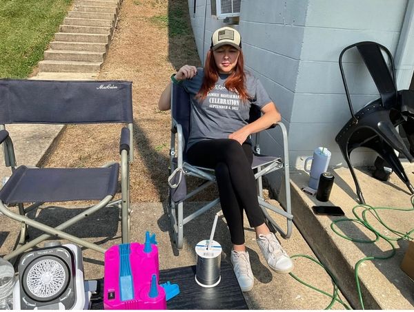 Woman relaxing outdoors on a chair with eyes closed, surrounded by chairs and outdoor equipment.