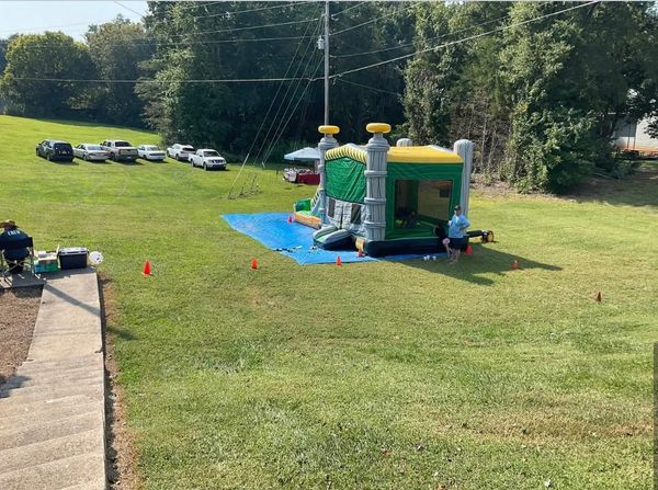 A bounce house setup on a grassy field with a few people and parked cars nearby.