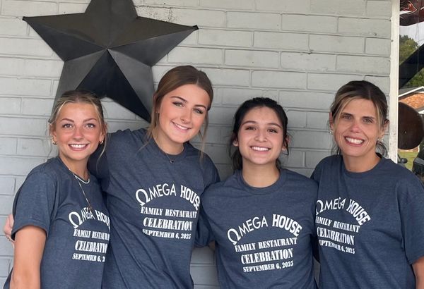 Four smiling women in matching Omega House celebration shirts pose in front of a brick wall with a star decoration.