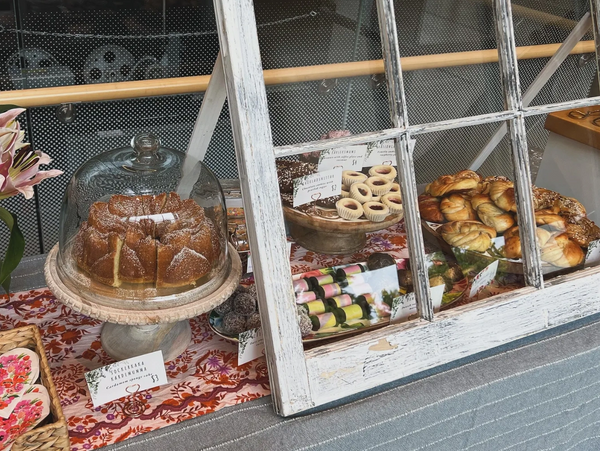 Rustic bakery display with cakes, pastries, and wine bottles behind a vintage window frame.