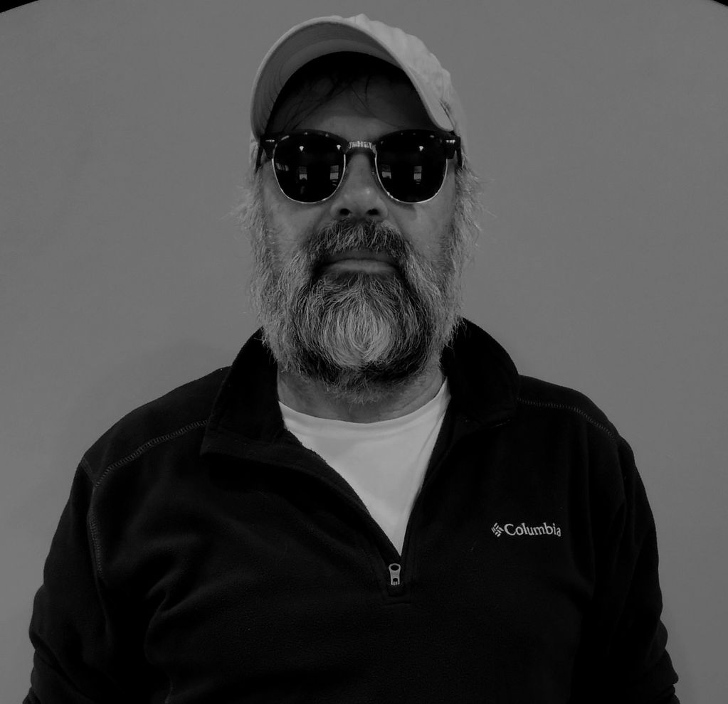 Bearded man wearing sunglasses and a cap in a black and white photo.