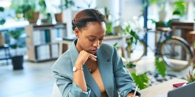 A woman in a blue blazer writing thoughtfully at a desk in a bright office.