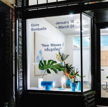 Art exhibit window display with plants and blue decor titled "New Waves" by Elvire Bonduelle.
