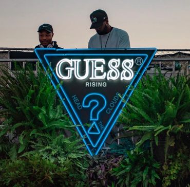 Two men behind a neon Guess Rising sign surrounded by greenery.