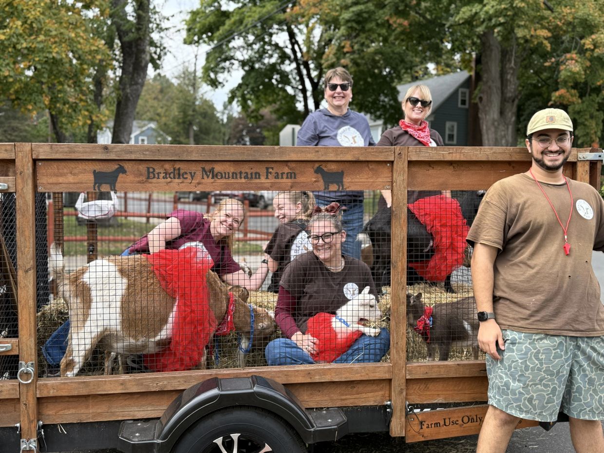 People smiling with goats dressed in red accessories inside a Bradley Mountain Farm trailer.