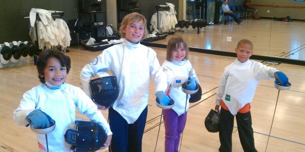 Four children in fencing gear holding foils and masks, smiling in a training room.