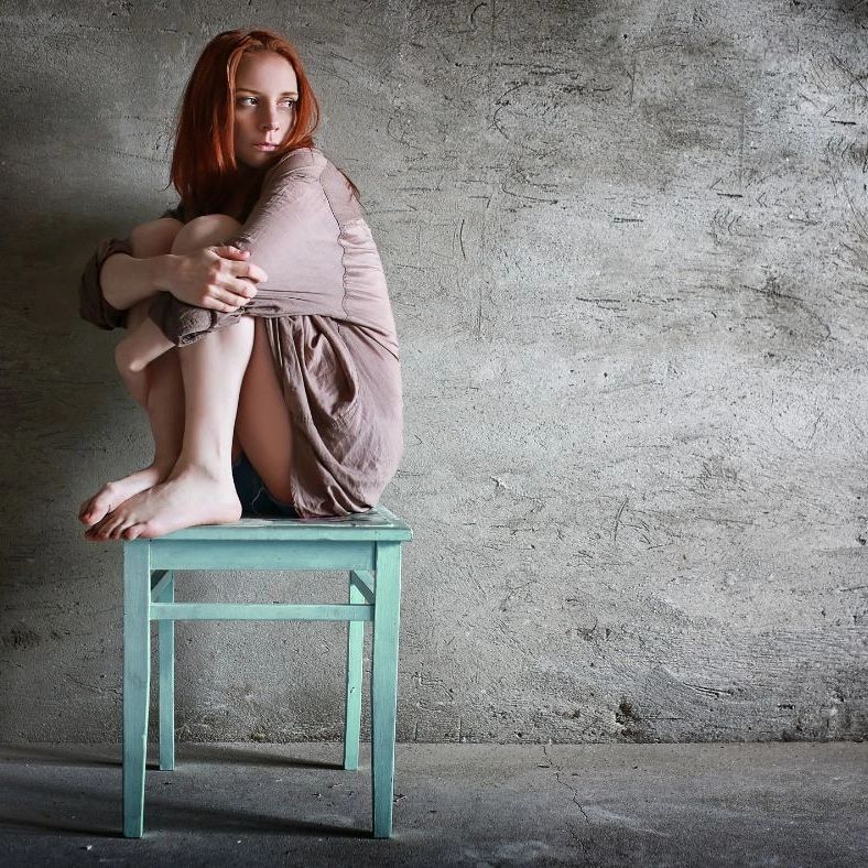 A woman with red hair sits curled up on a teal chair against a rough concrete wall.