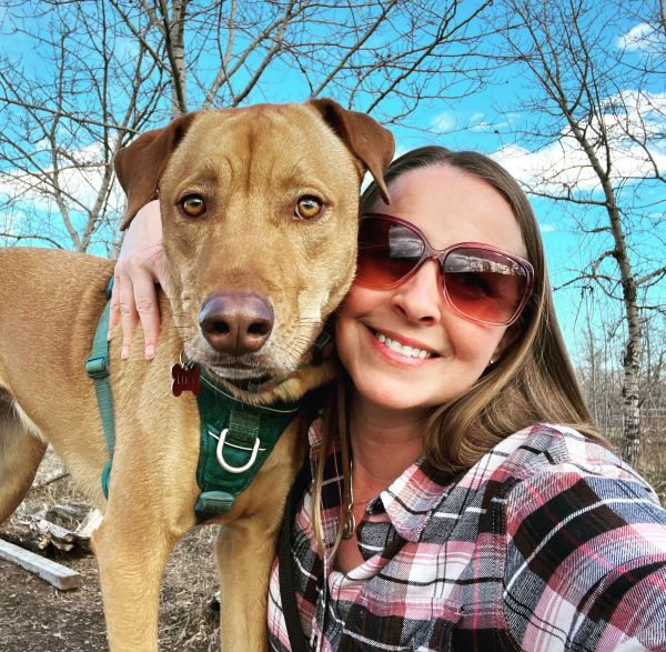 Woman with sunglasses takes a selfie hugging her large brown dog outdoors.