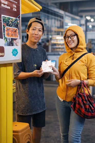 Two people smiling and exchanging a small wrapped item at a market stall.