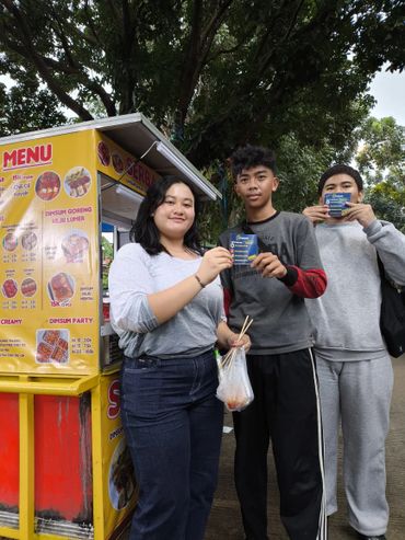 Three young people holding cards near a street food stall under a tree.