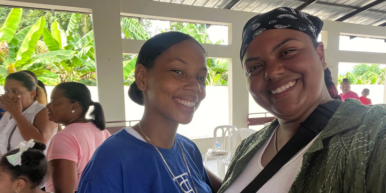 Two smiling women posing for a selfie in a bright, open space with greenery outside.