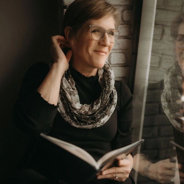 Woman in glasses reading a book by a window, smiling thoughtfully.