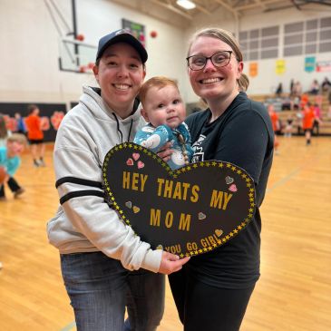 Two women and a baby hold a heart-shaped sign at a basketball gym.