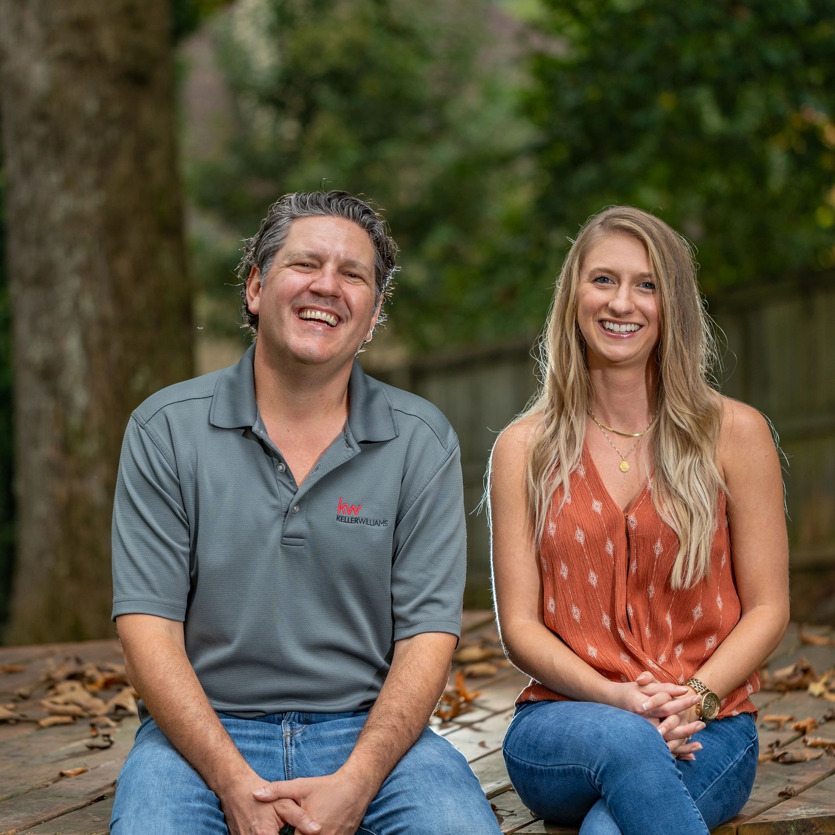 Smiling man and woman sitting outdoors with trees in the background.