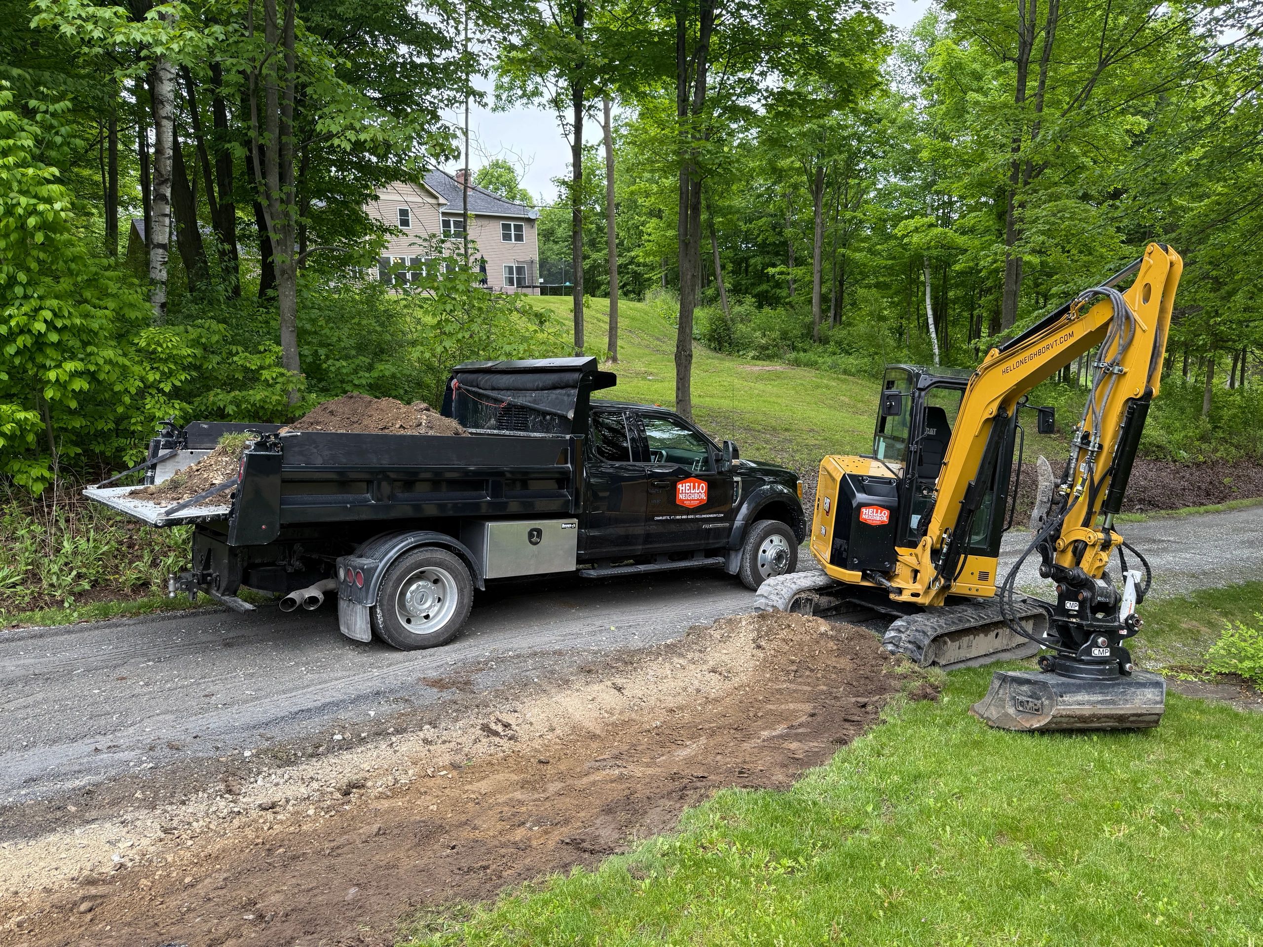 Black dump truck and yellow excavator at a roadside construction site beside a green wooded area.