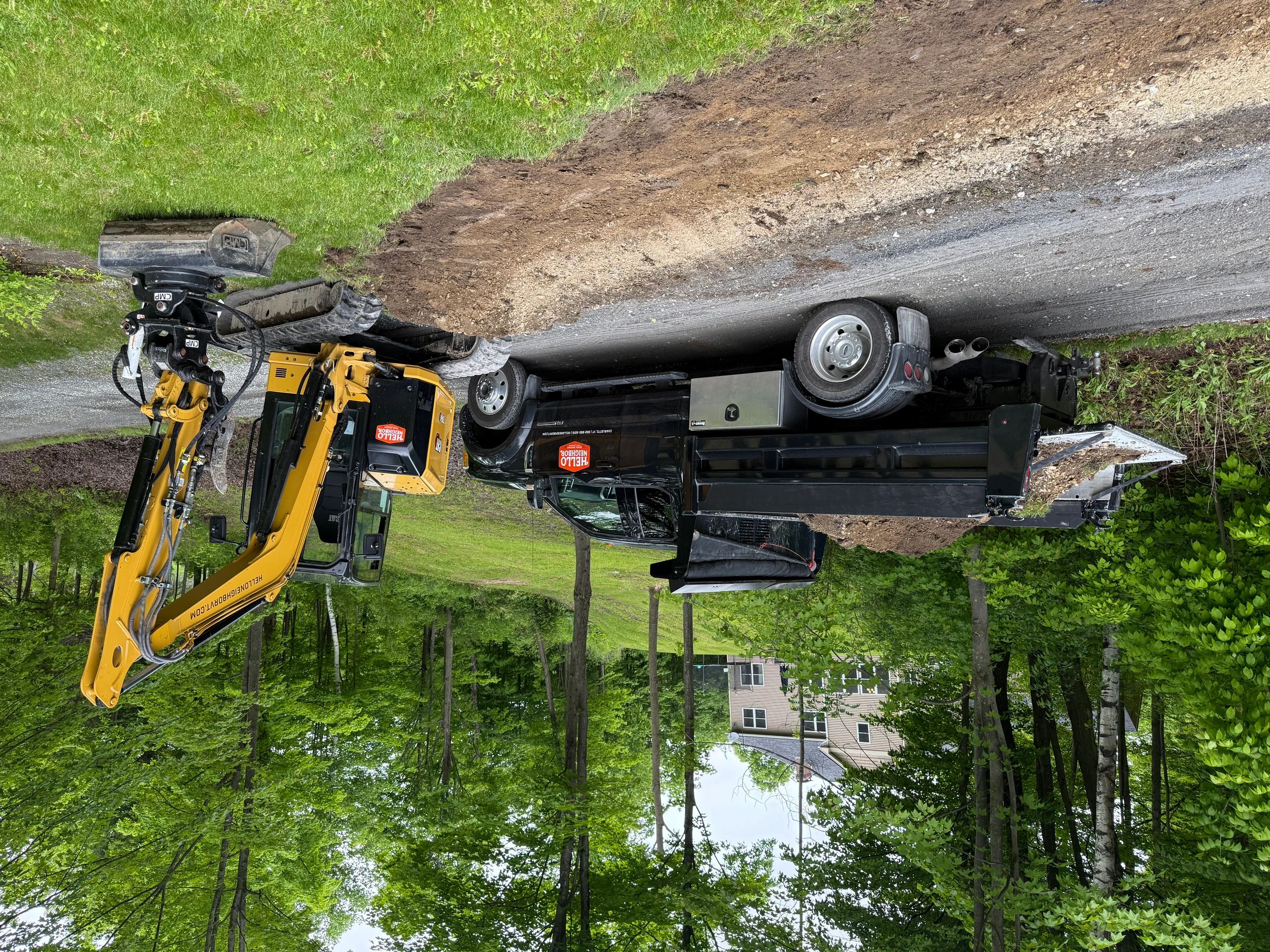 Black dump truck and yellow excavator at a roadside construction site beside a green wooded area.