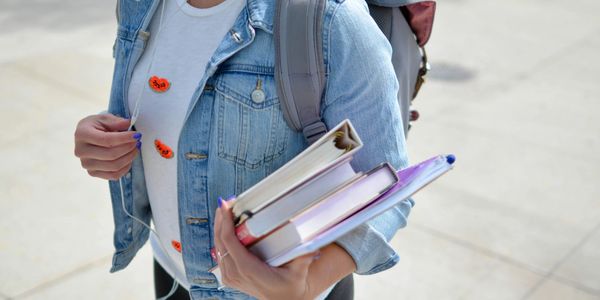 Student in denim jacket holding books and wearing backpack outdoors.