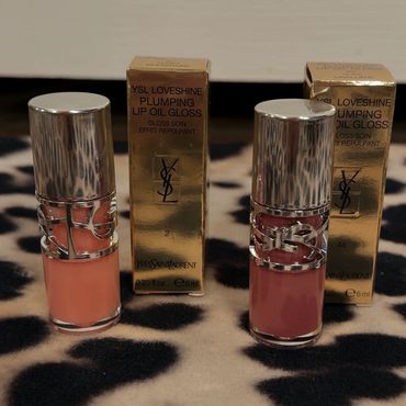 Two YSL Plumping Lip Oil Gloss bottles with gold packaging on a leopard print fabric.