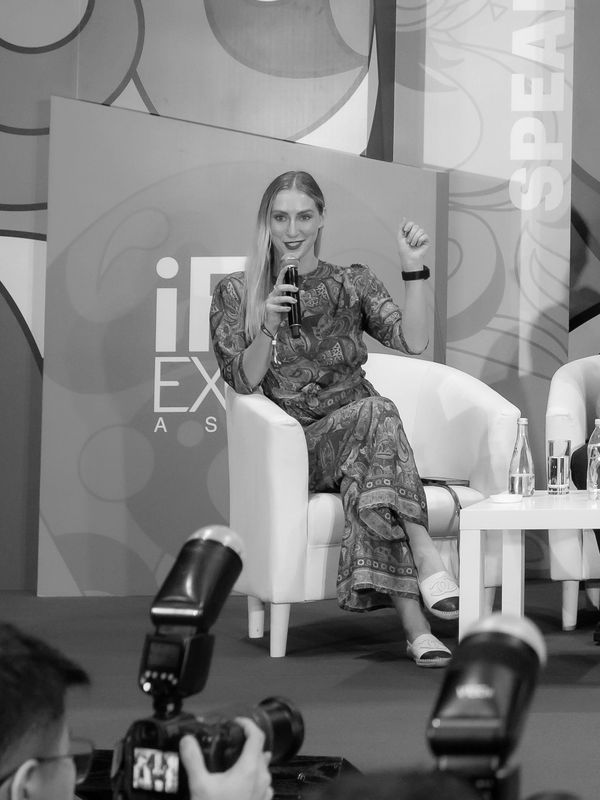 Woman speaking into microphone at a panel event, seated in a white armchair.
