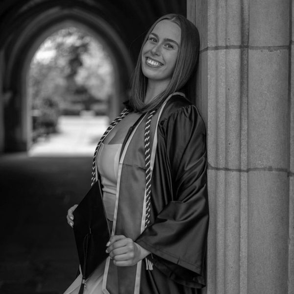Smiling graduate in cap and gown leans against a stone pillar.