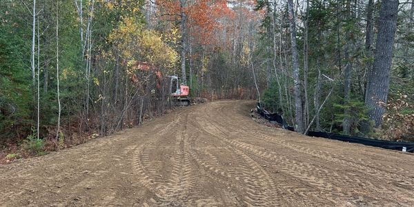 A dirt road under construction curves through a forest with autumn foliage.