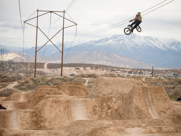 A BMX rider performs a high jump over dirt ramps with mountains in the background.