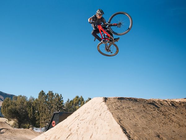 A biker performs an aerial trick over a dirt jump against a clear blue sky.