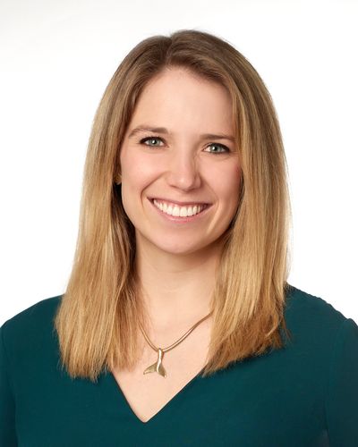 Smiling woman with blonde hair wearing a teal top and whale tail necklace.
