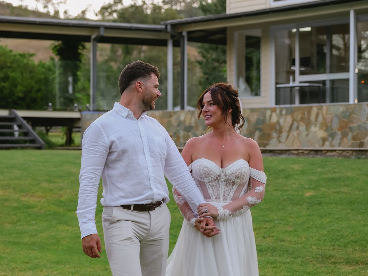 Newlyweds walk hand in hand on green lawn, smiling at each other.