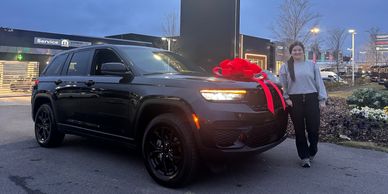 Woman standing next to a black SUV with a red bow in a dealership lot at dusk.