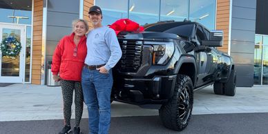 A happy couple stands beside a new black truck with a big red bow.