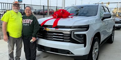 Couple standing next to a new white SUV with a large red bow on the hood.